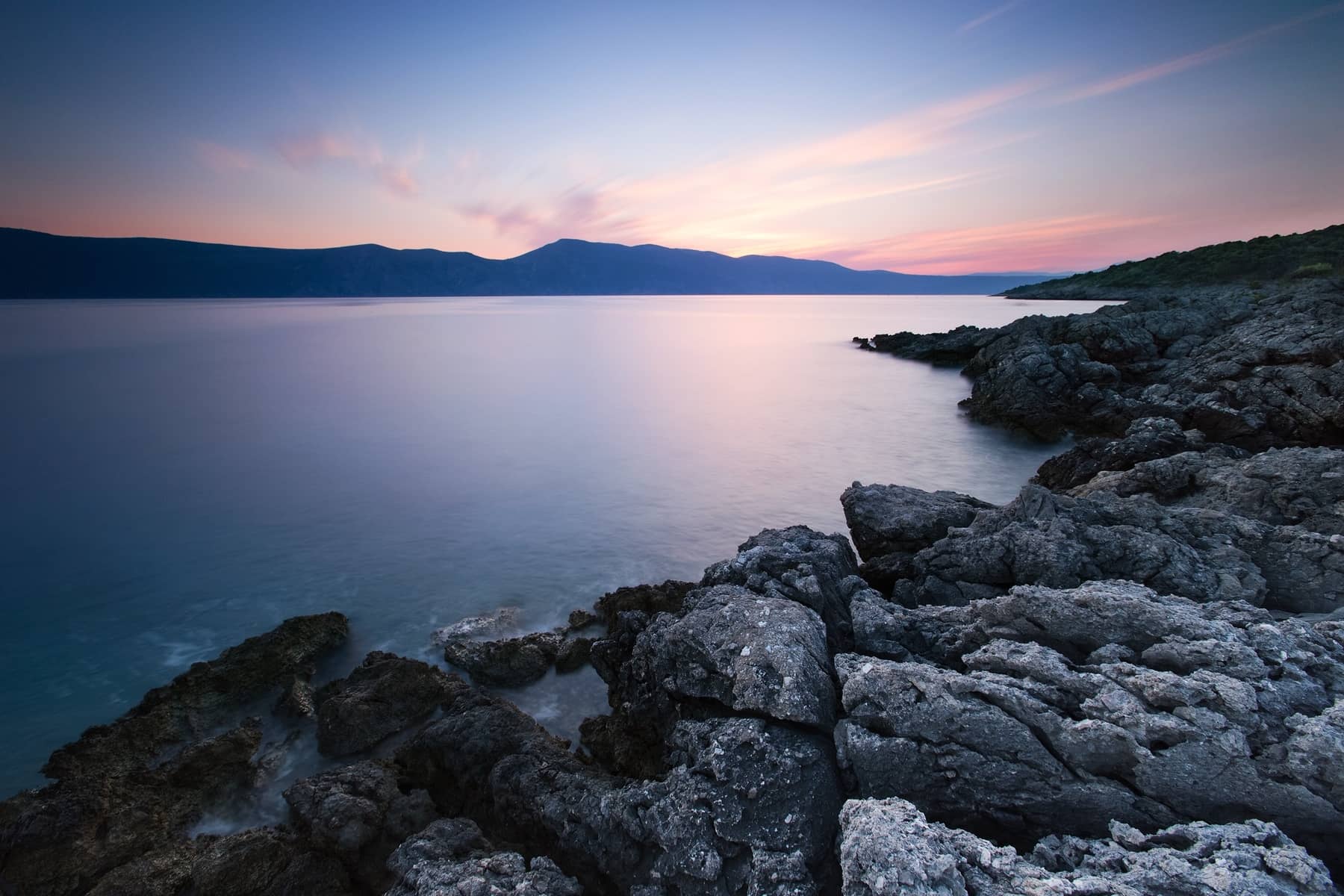 Peaceful rocky beach at sunrise 
                        with the sun turning the sky pink from behind the mountains in the distance